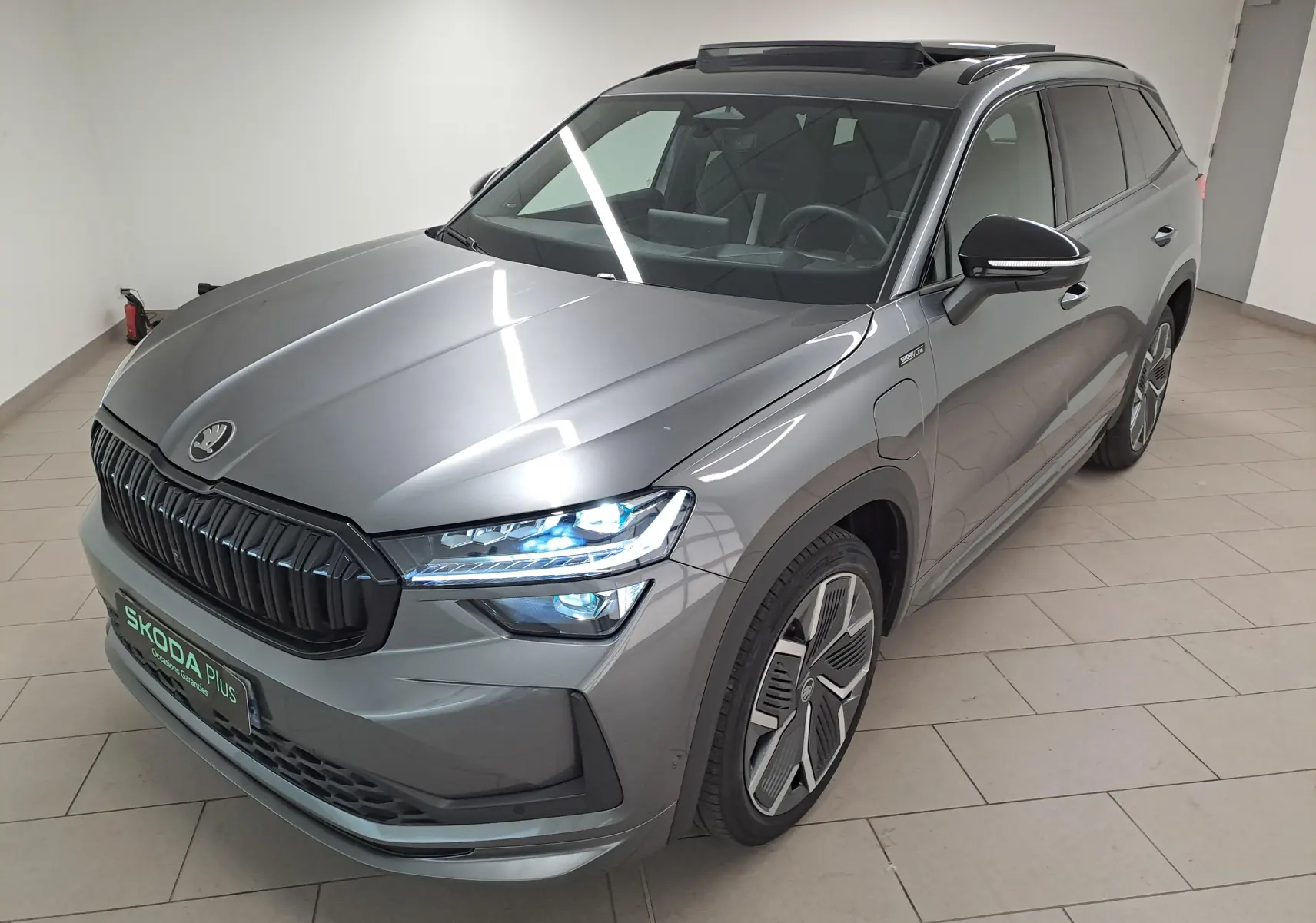 Vue 3/4 avant droite du Skoda Kodiaq gris graphite avec phares allumés et toit panoramique ouvrant visible.