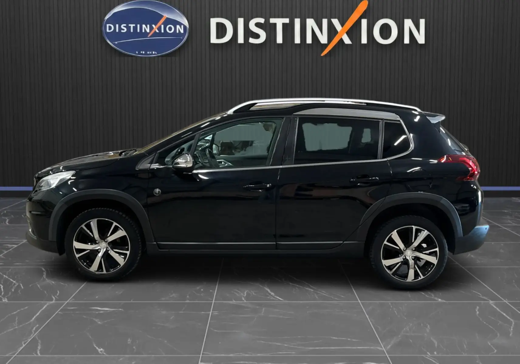 Profil droit d'un Peugeot 2008 noir 2018 avec jantes alliage et barres de toit, sur fond intérieur avec logo Distinxion.