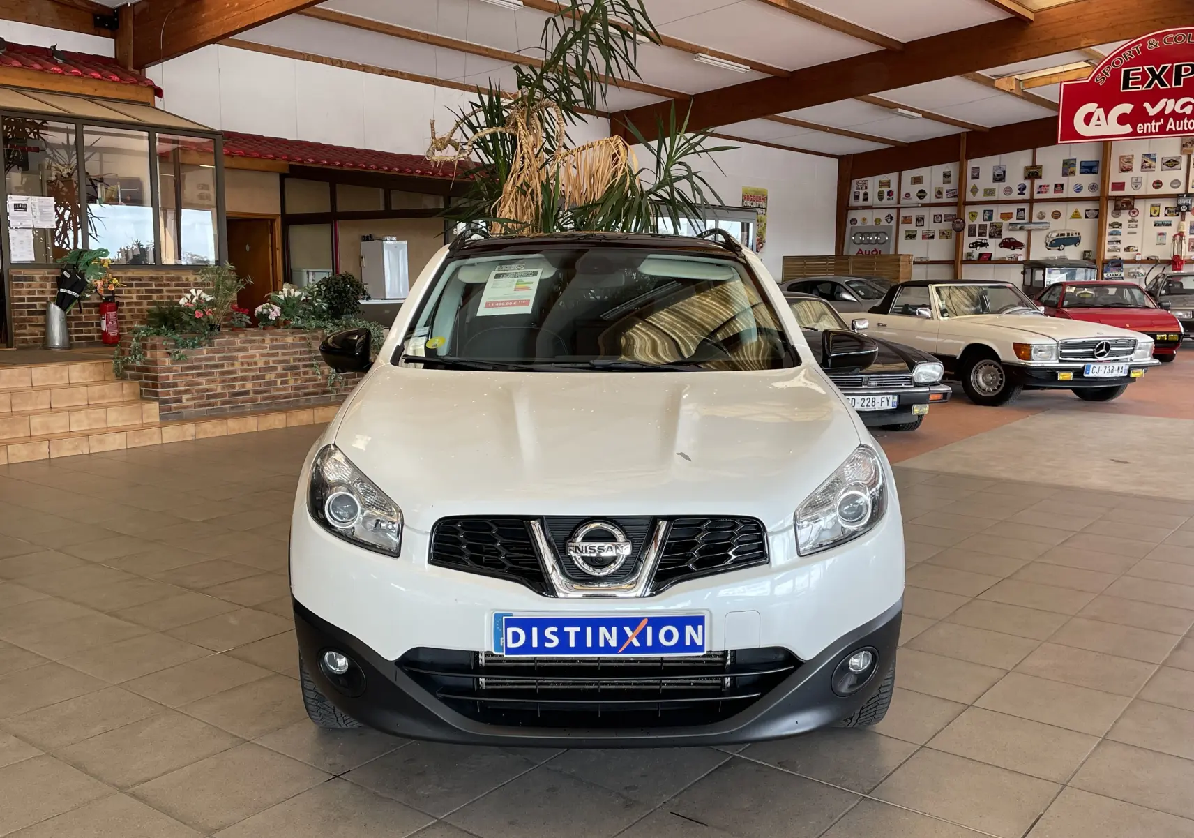 Nissan Qashqai blanc en vue frontale, intérieur showroom avec plusieurs voitures classiques en arrière-plan.