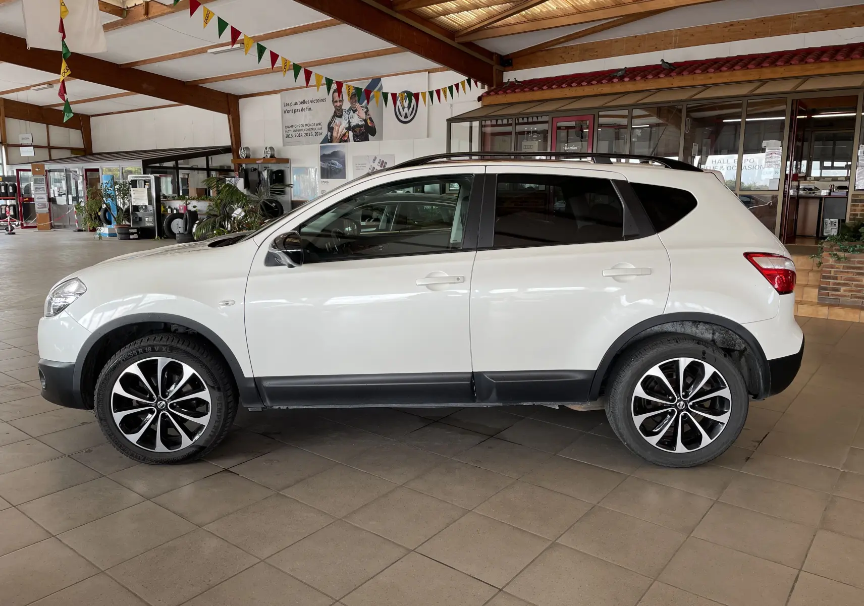 Profil côté gauche d'un Nissan Qashqai blanc 2013 avec jantes noires et blanches, garé en intérieur lumineux.