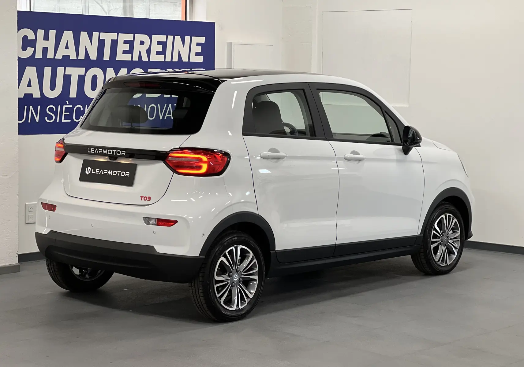 Vue 3/4 arrière droite d'une Leapmotor T03 blanche avec toit noir et feux arrière allumés en intérieur showroom.