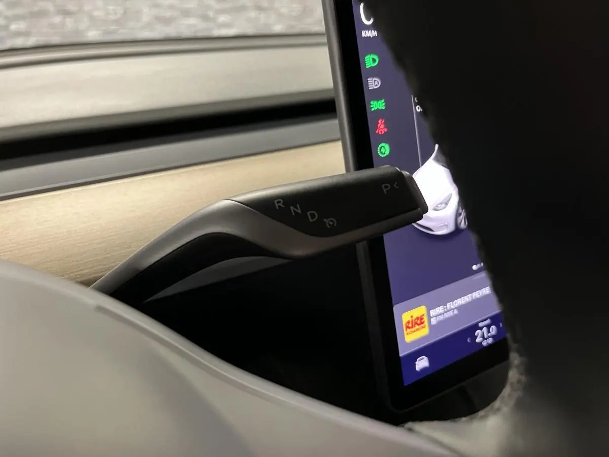 Gros plan sur la commande de boîte automatique noire et argentée, avec l'écran tactile et le tableau de bord bois clair d'une Tesla Model Y blanche.
