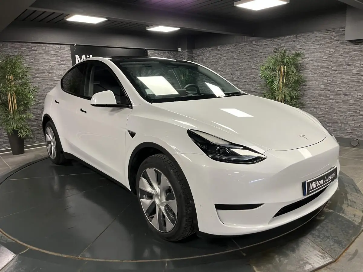 Tesla Model Y blanc nacré vu en 3/4 avant droit dans un showroom avec toit panoramique en verre visible.