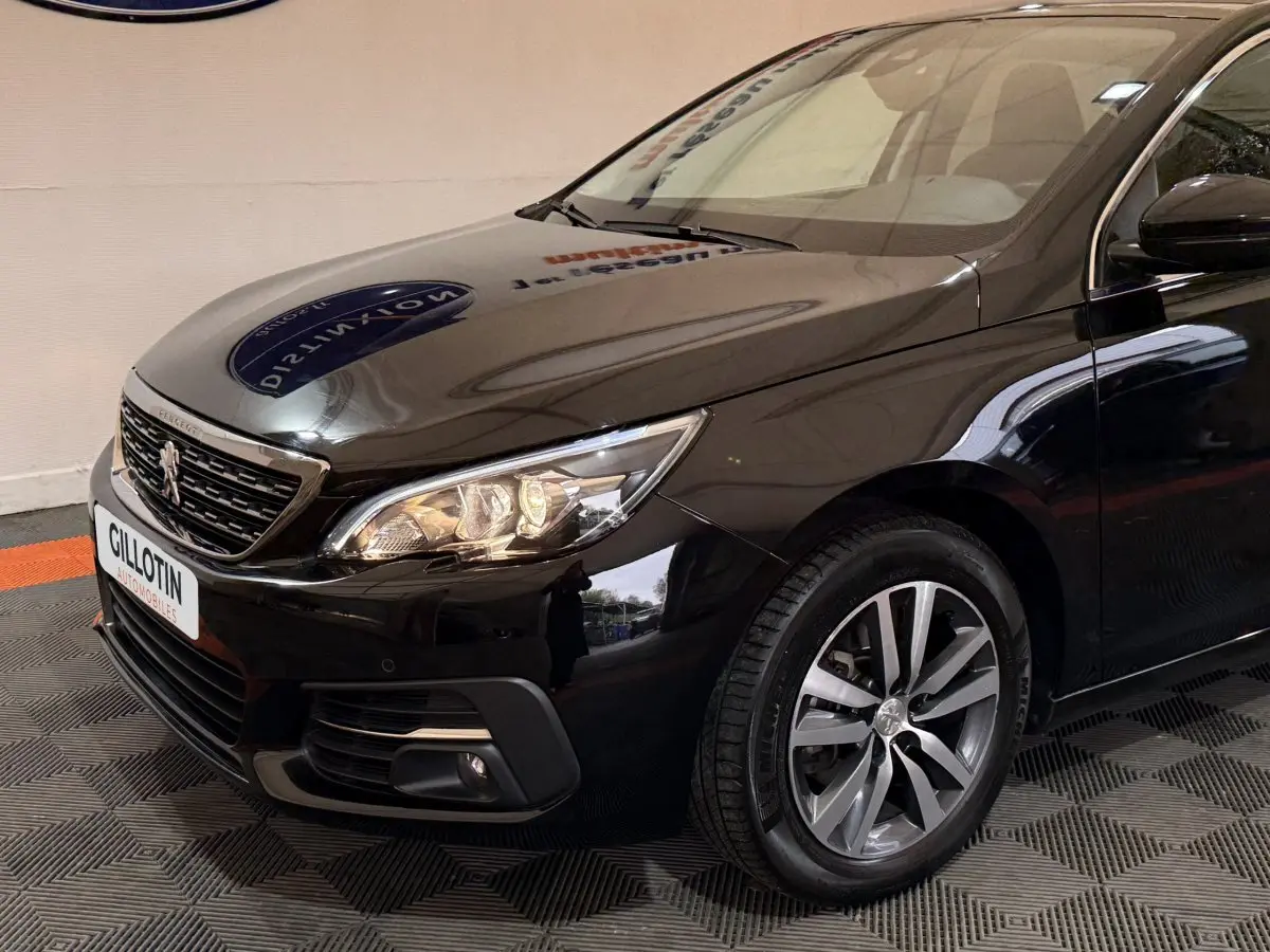 Vue 3/4 avant droit d'une Peugeot 308 noire avec phares allumés et jantes alliage visibles.