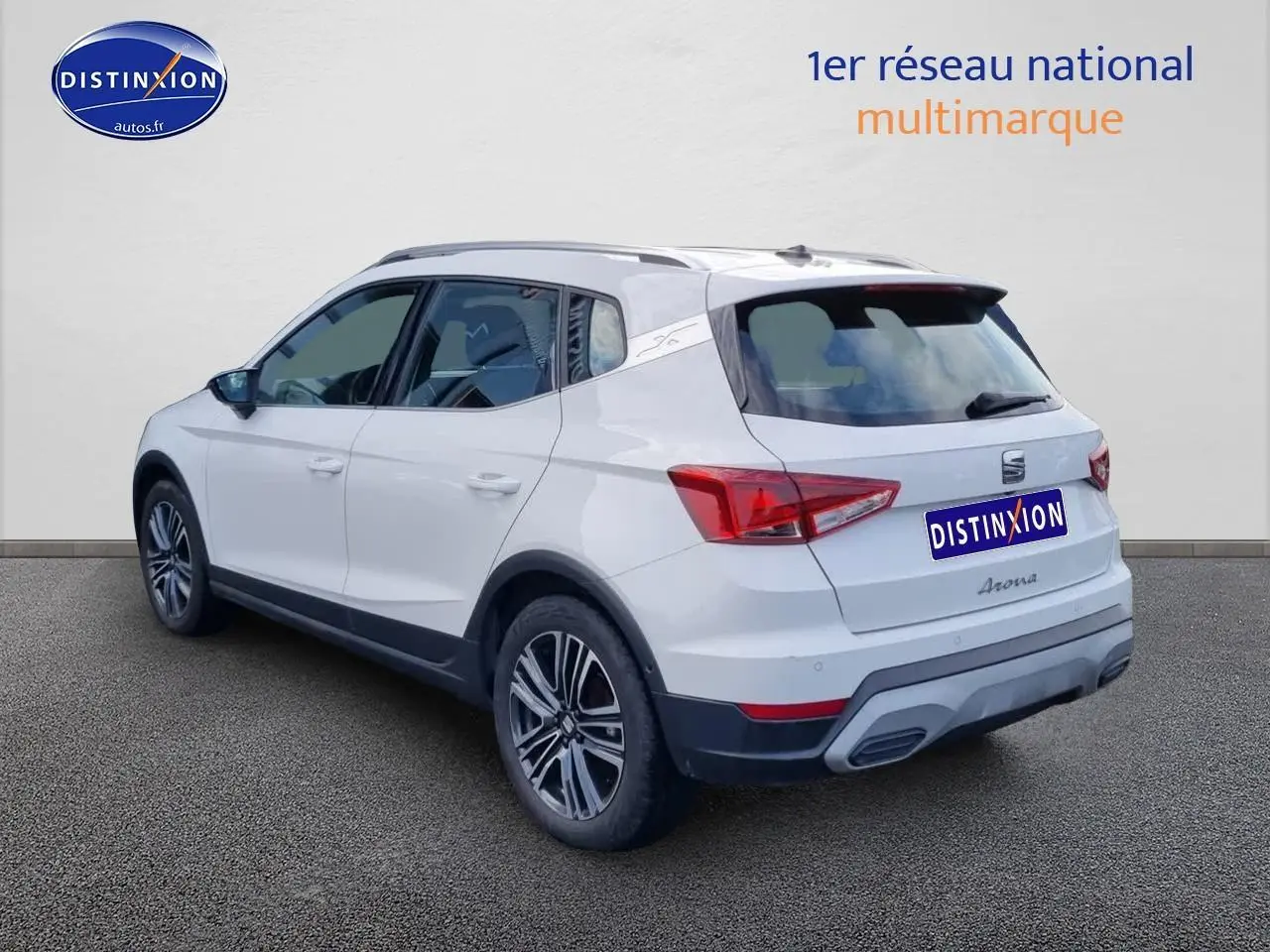 Vue 3/4 arrière droite d'un SEAT Arona blanc 2023 avec jantes alu et barres de toit visibles.