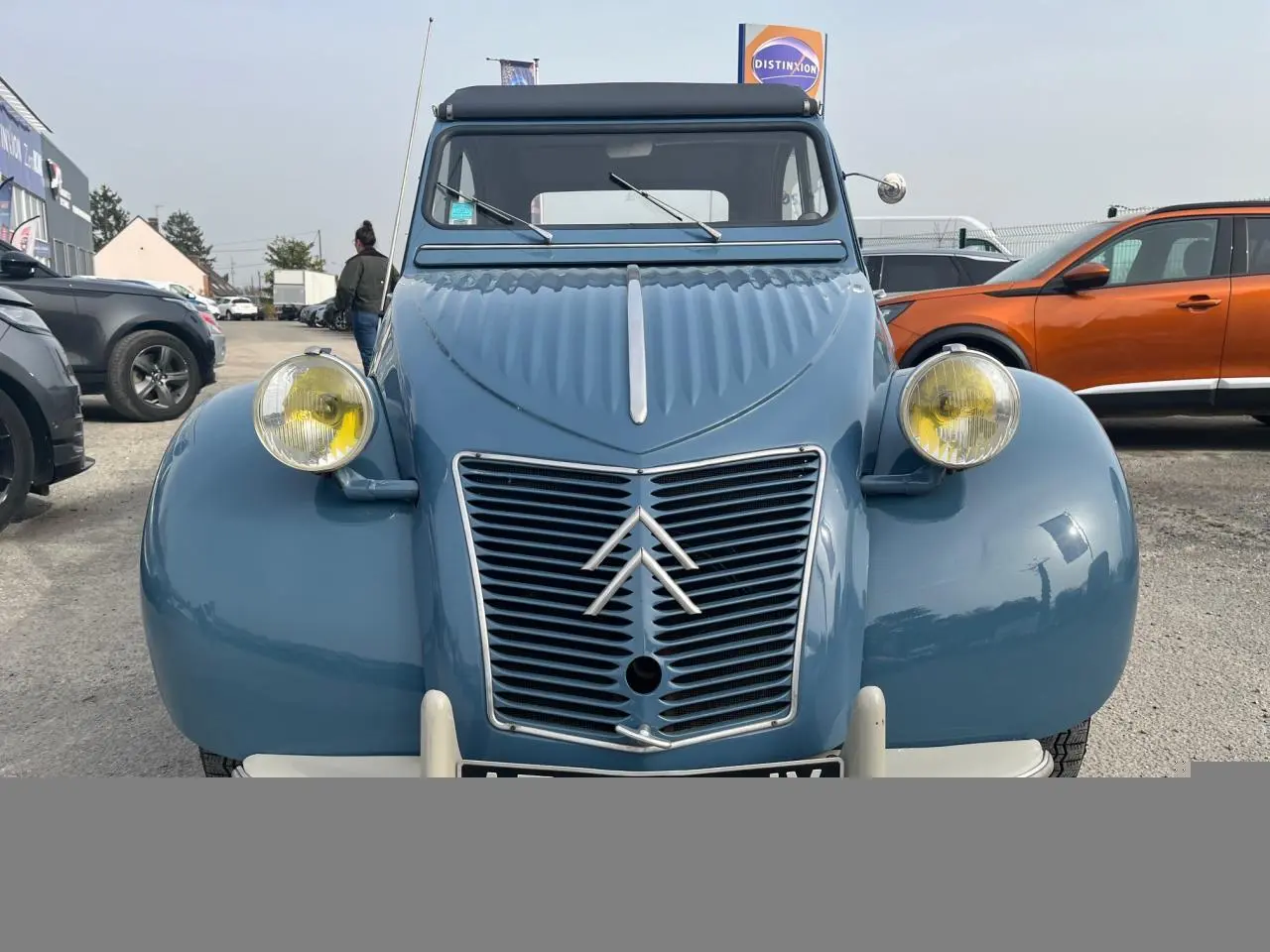 Vue frontale d'une Citroën 2 CV bleu clair de 1960 avec phares ronds jaunes et calandre à chevrons emblématiques.