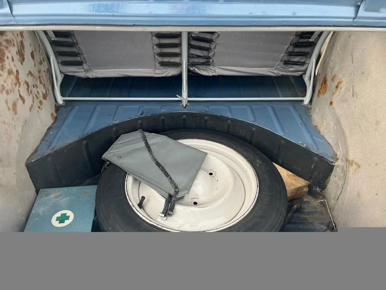 Vue intérieure du coffre d'une Citroën 2 CV bleu clair de 1960, avec roue de secours et trousse à outils grise.