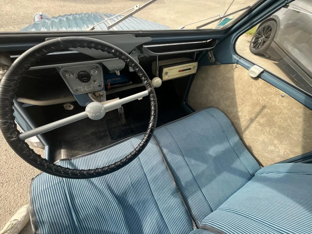Vue intérieure du poste de conduite d'une Citroën 2 CV 1960 bleu clair, avec volant fin et sièges rayés bleus.