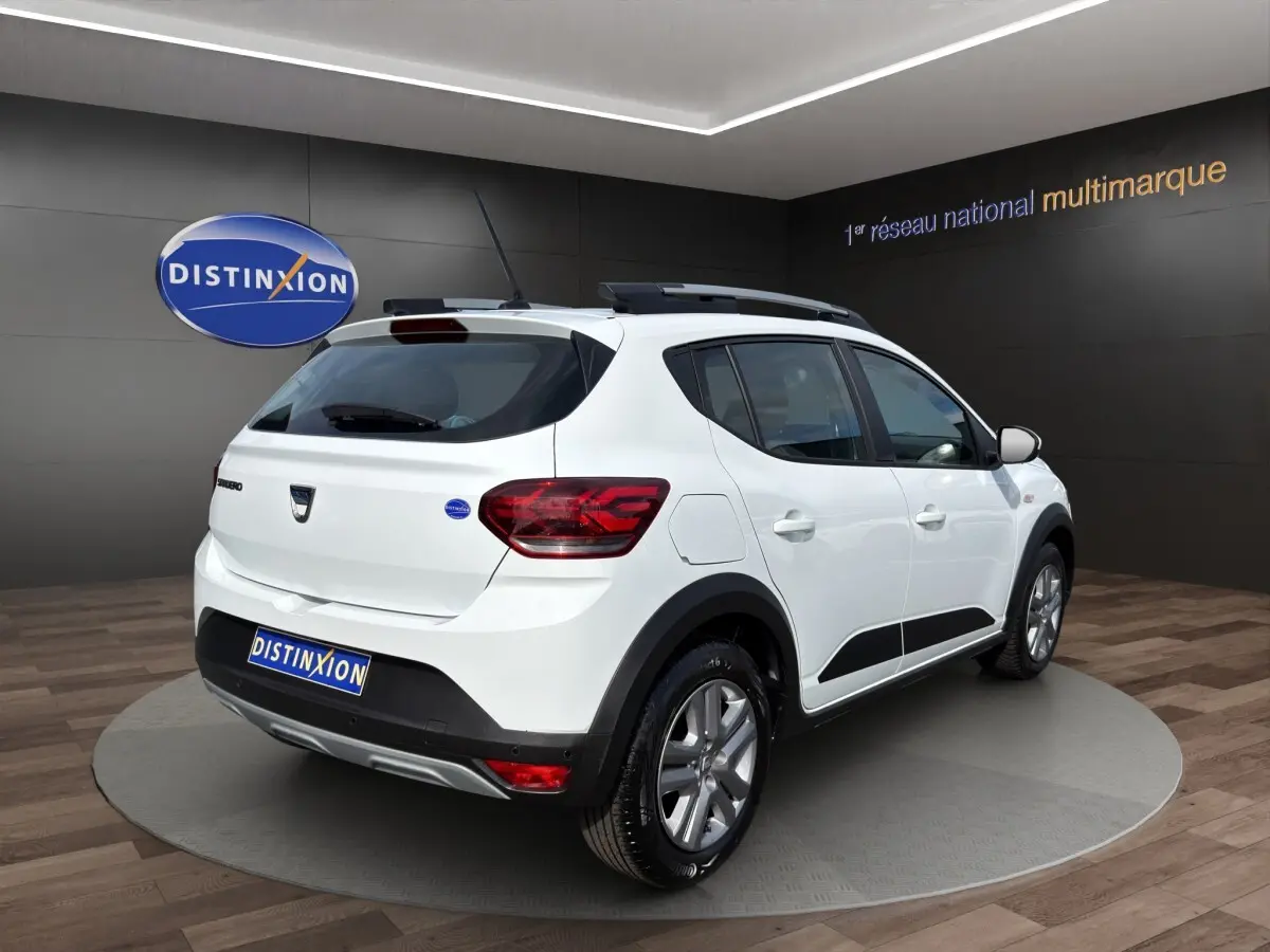 Vue 3/4 arrière droite d'une Dacia Sandero Stepway blanche avec barres de toit et protections noires visibles.
