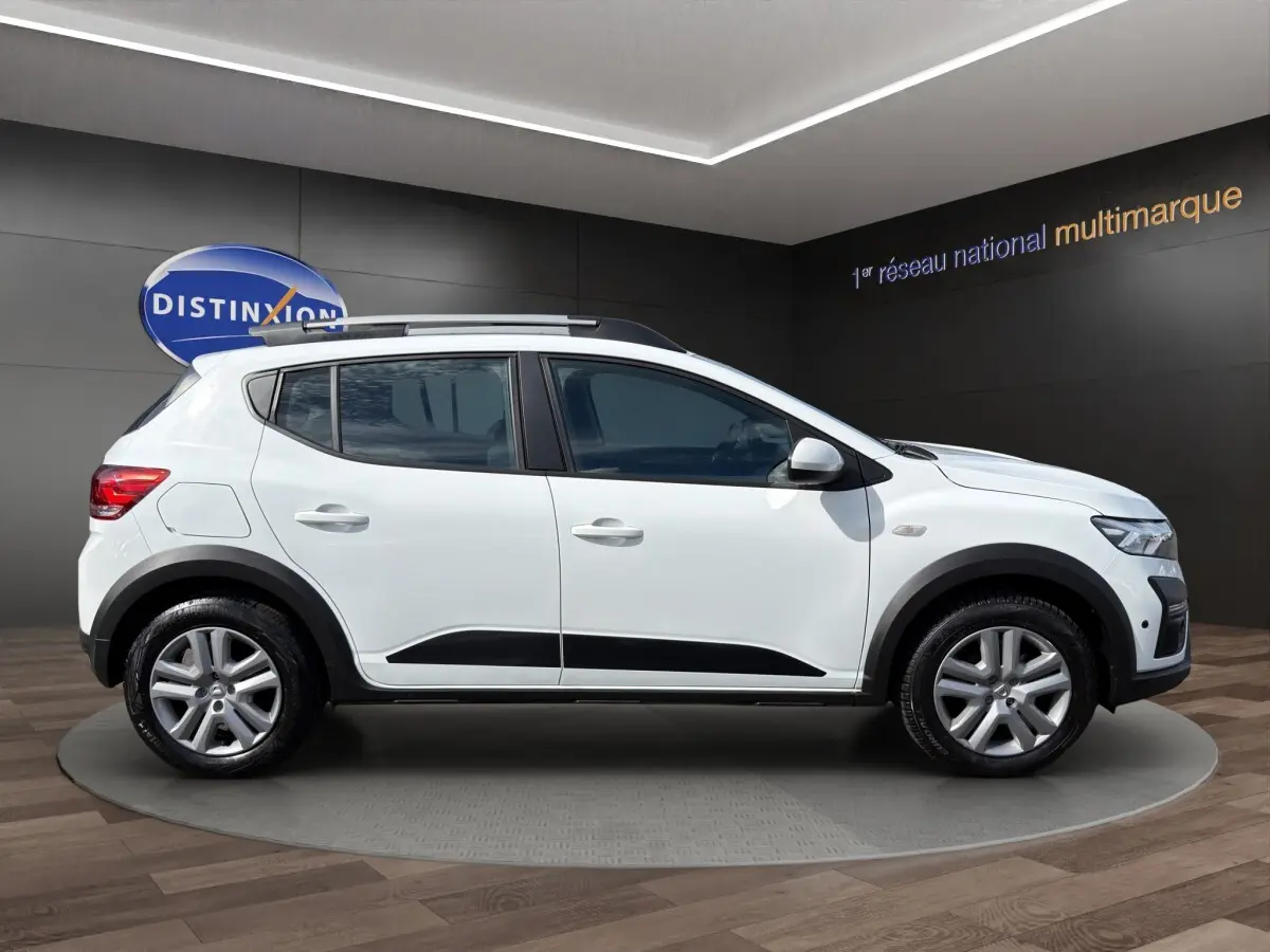 Profil droit de la Dacia Sandero Stepway blanche 2022 avec protections noires et barres de toit visibles en intérieur showroom.