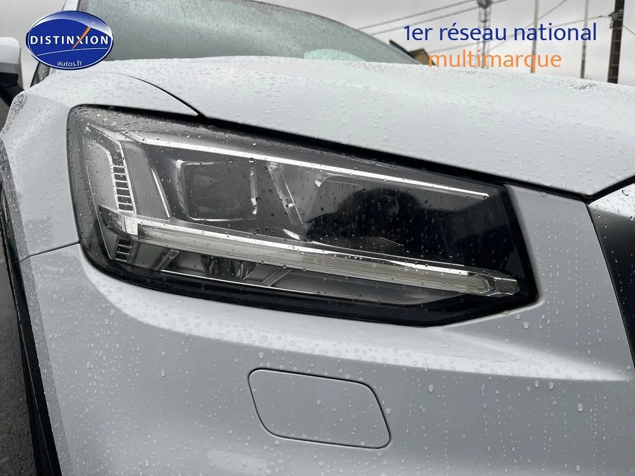 Gros plan sur le phare avant droit d’un Audi Q2 blanc Ibis avec gouttes de pluie visibles sur la carrosserie.