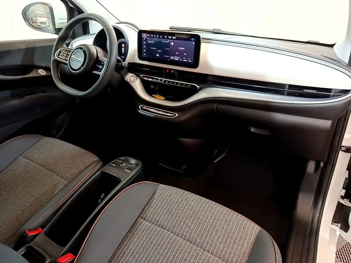 Intérieur avant de la Fiat 500 électrique blanche, tableau de bord moderne avec écran tactile central et sièges gris à surpiqûres rouges.
