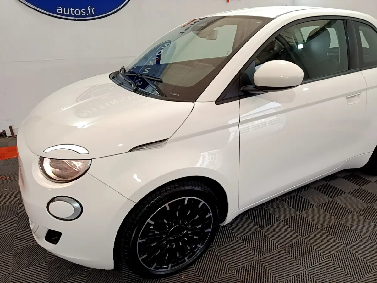 Vue 3/4 avant droit d'une Fiat 500 électrique blanche avec jantes noires et phares allumés.