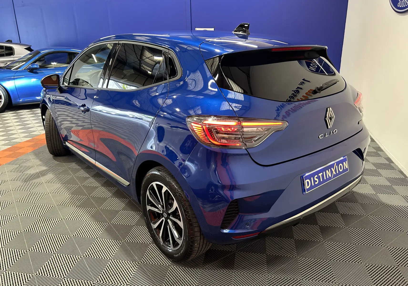 Vue 3/4 arrière droite d'une Renault Clio V bleu Iron Metal avec feux arrière allumés dans un showroom.