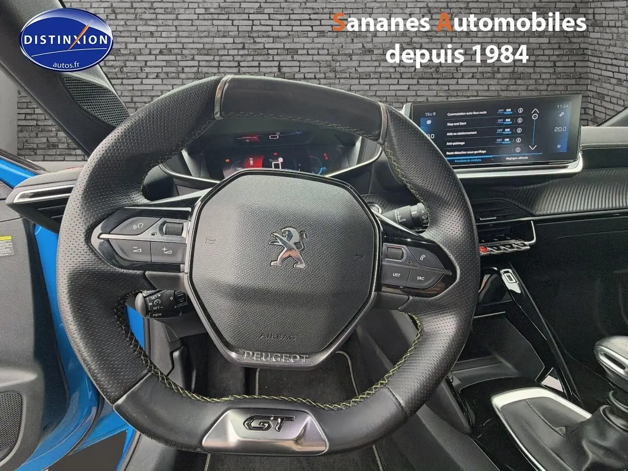 Vue centrée sur le volant cuir noir GT avec coutures vertes, tableau de bord numérique et écran tactile du Peugeot 208 bleu clair.