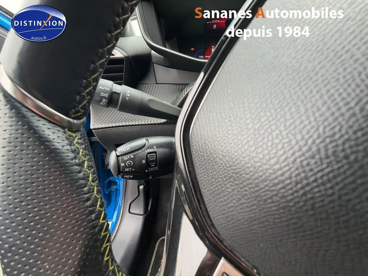 Vue rapprochée du volant cuir noir avec surpiqûres vertes et commandes, tableau de bord partiel d'une Peugeot 208 bleu clair 2020.