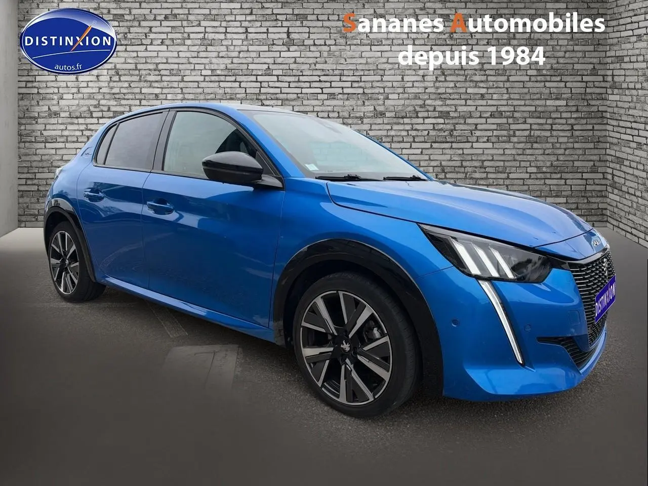 Peugeot 208 bleu clair en 3/4 avant droit, avec jantes alliage et feux LED distinctifs, sur fond mur en briques.