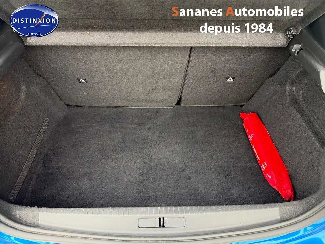 Coffre ouvert d'une Peugeot 208 GT Line bleu clair, montrant un espace de rangement propre avec un triangle de signalisation rouge.