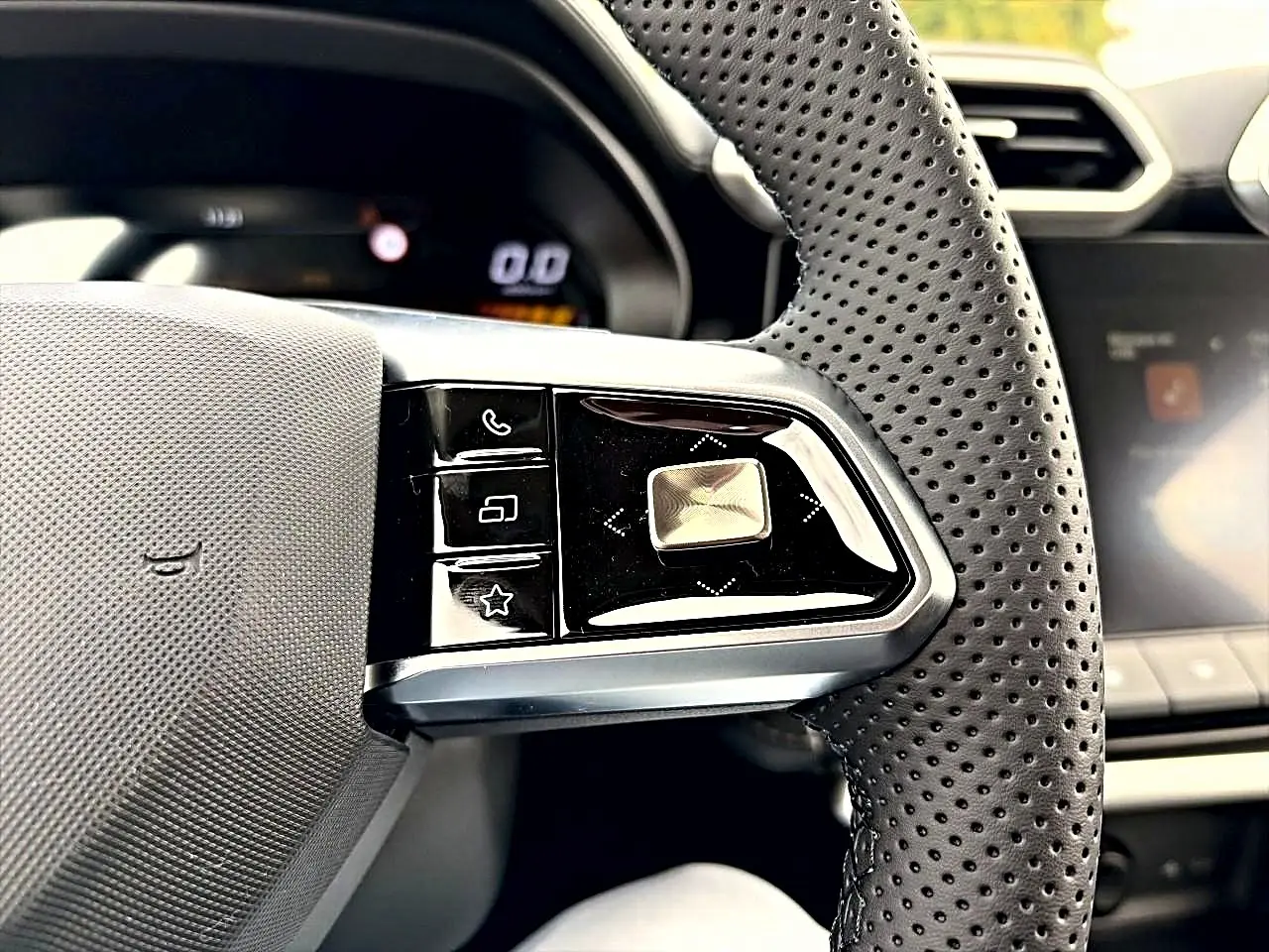 Gros plan sur les commandes gauche du volant perforé noir du MG MOTOR ZS 1.5 hybrid Camden grey, tableau de bord flou en arrière-plan.
