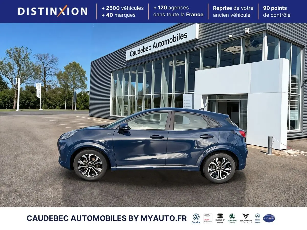 Ford Puma bleu en profil droit, avec jantes alliage 17 pouces et arches couleur carrosserie, devant un showroom.