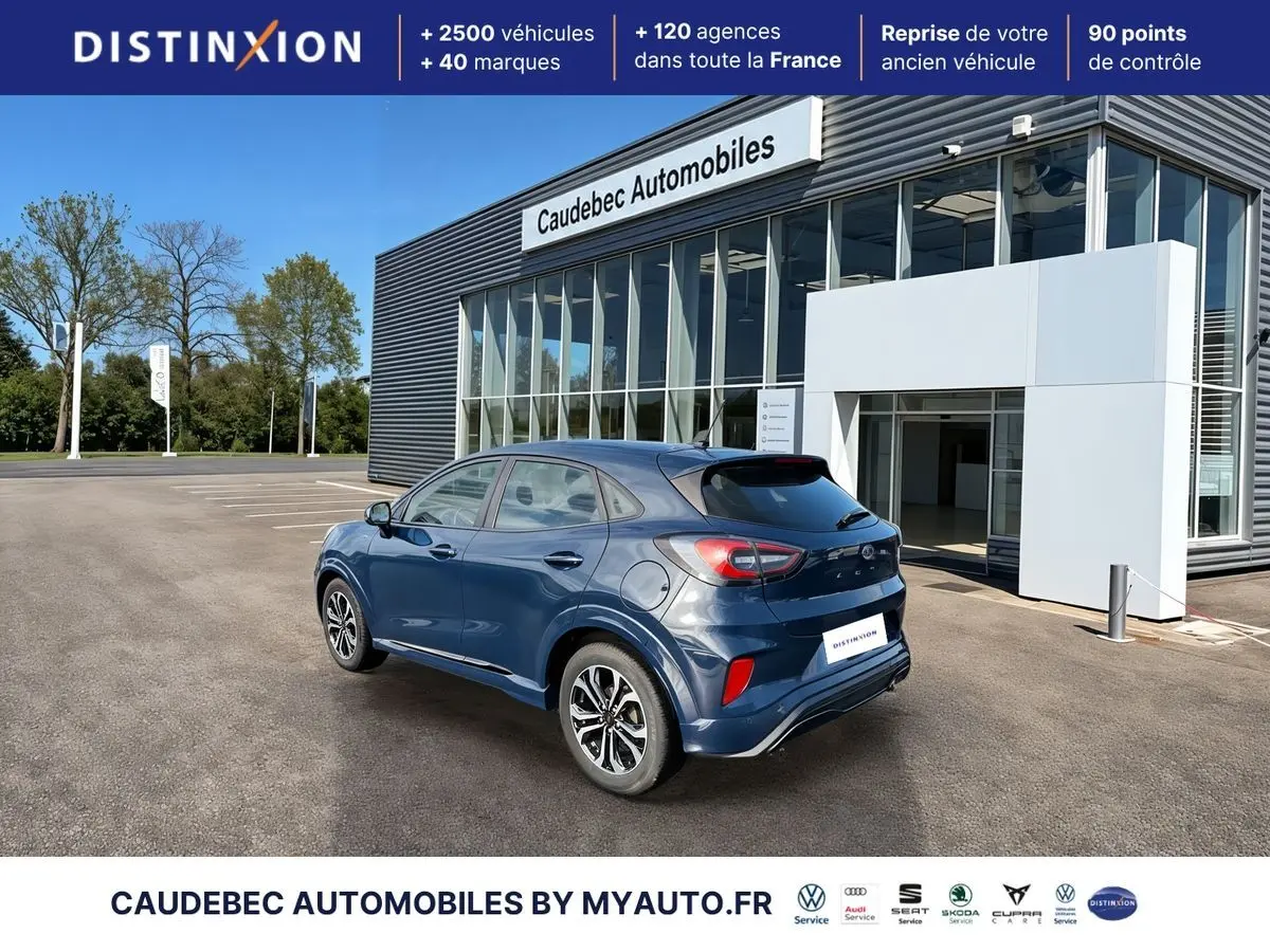 Ford Puma bleu en 3/4 arrière droit, avec jantes alliage 17 pouces et feux arrière LED visibles, devant un showroom.