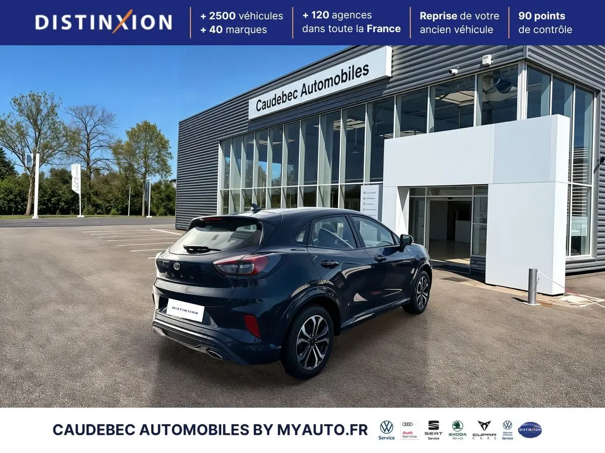 Vue 3/4 arrière droite d’un Ford Puma bleu métallisé avec jantes alliage 17 pouces devant un showroom moderne.