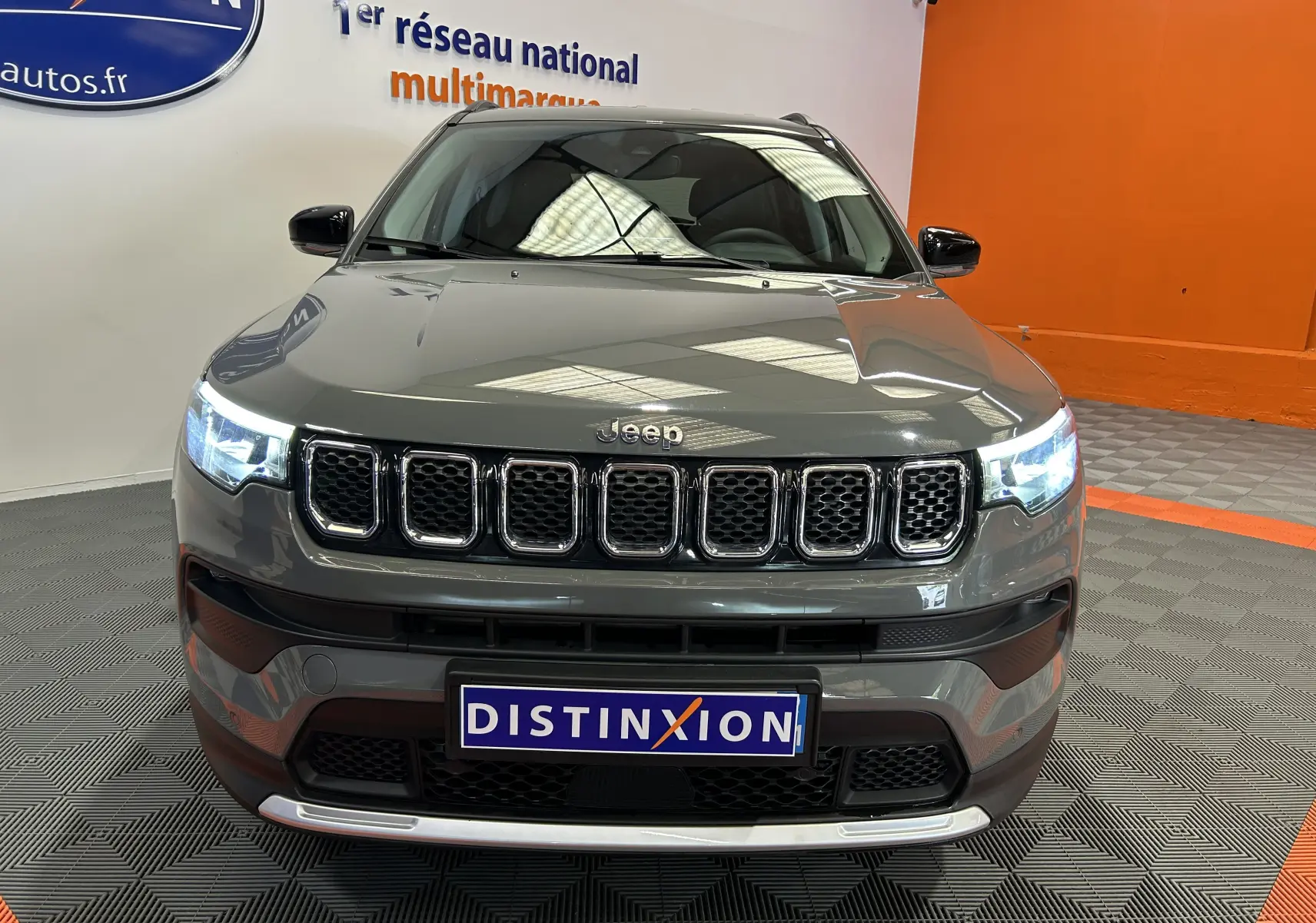 Vue frontale d'un Jeep Compass gris avec phares allumés et calandre à sept ouvertures, sur sol gris et fond orange.