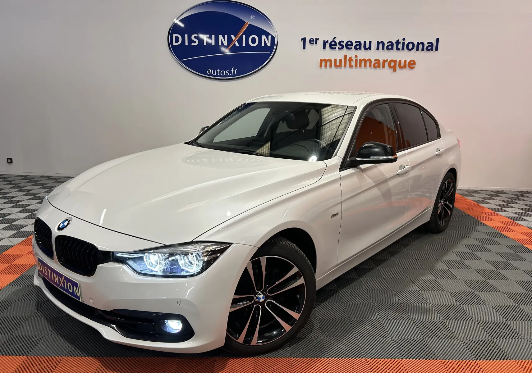 BMW Série 3 320i blanc vue 3/4 avant droit avec phares allumés et jantes noires bicolores dans un showroom.