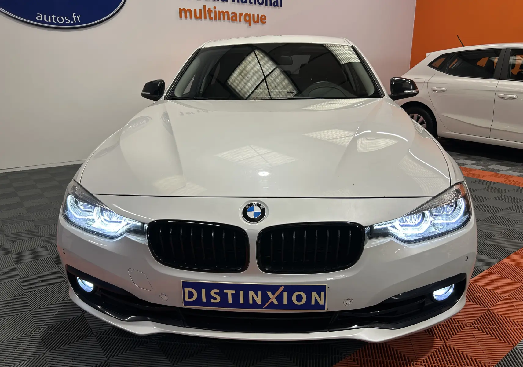 Vue frontale d'une BMW Série 3 320i blanche de 2018 avec phares LED allumés et calandre noire brillante.