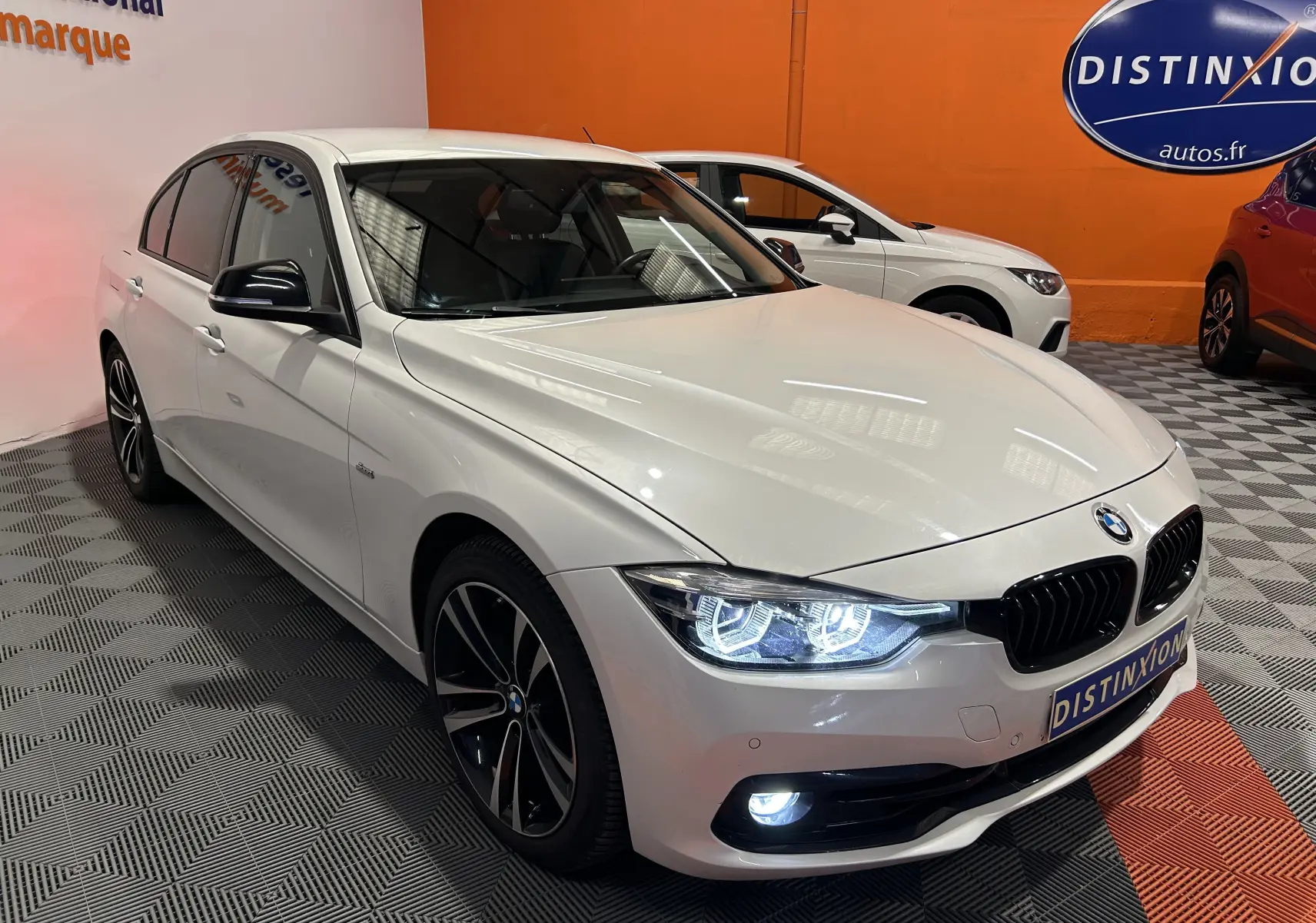 BMW Série 3 320i blanc vue 3/4 avant droit avec phares LED allumés et jantes sport bicolores.
