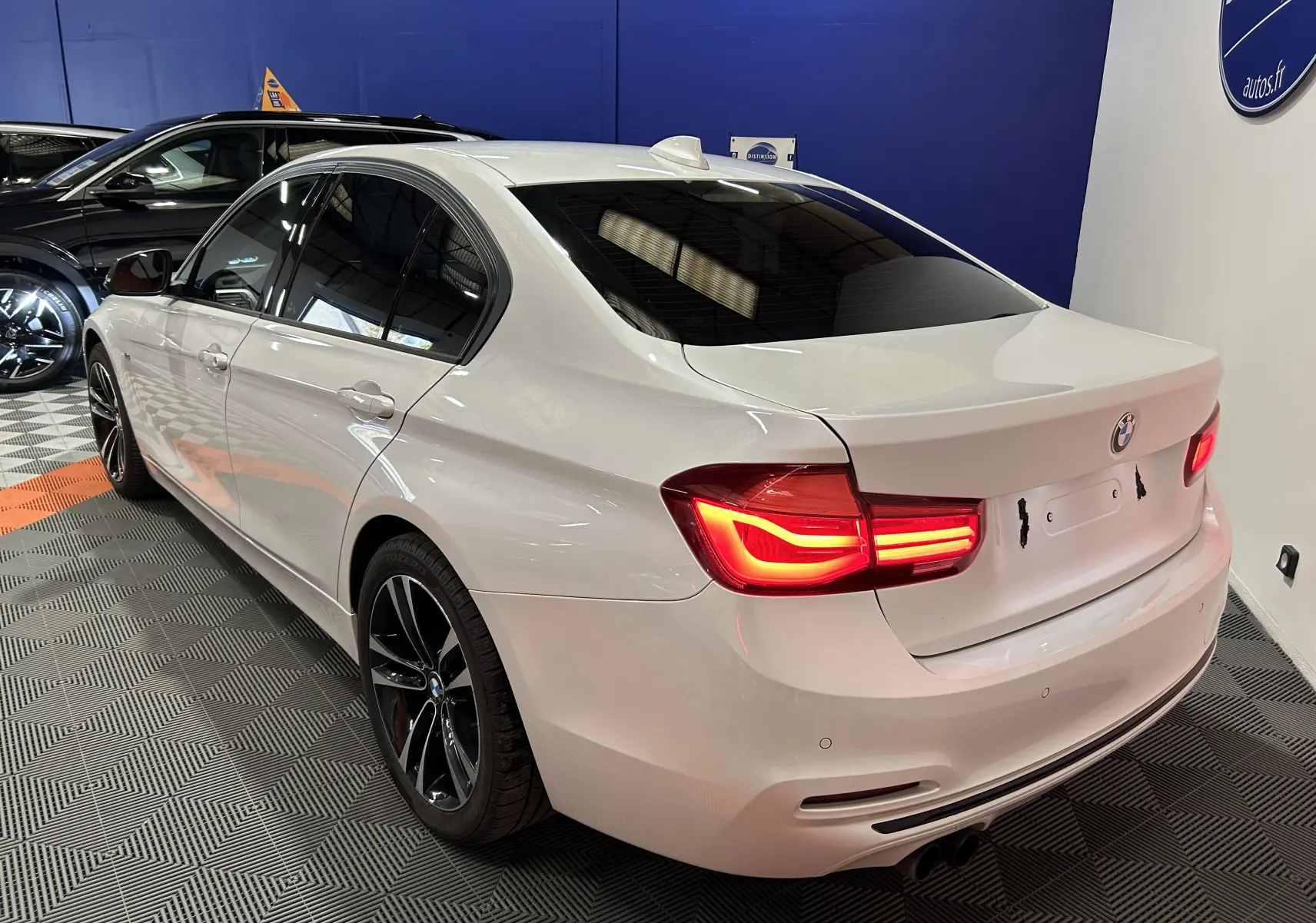 BMW Série 3 320i blanche vue 3/4 arrière droit avec feux allumés et jantes noires en intérieur showroom.
