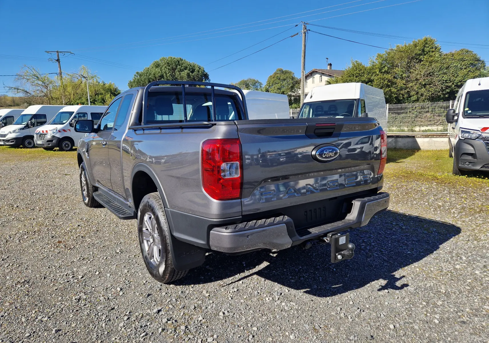 Vue 3/4 arrière droite d’un Ford Ranger Super Cabine gris carbone avec attelage et barres de protection sur un terrain gravillonné.