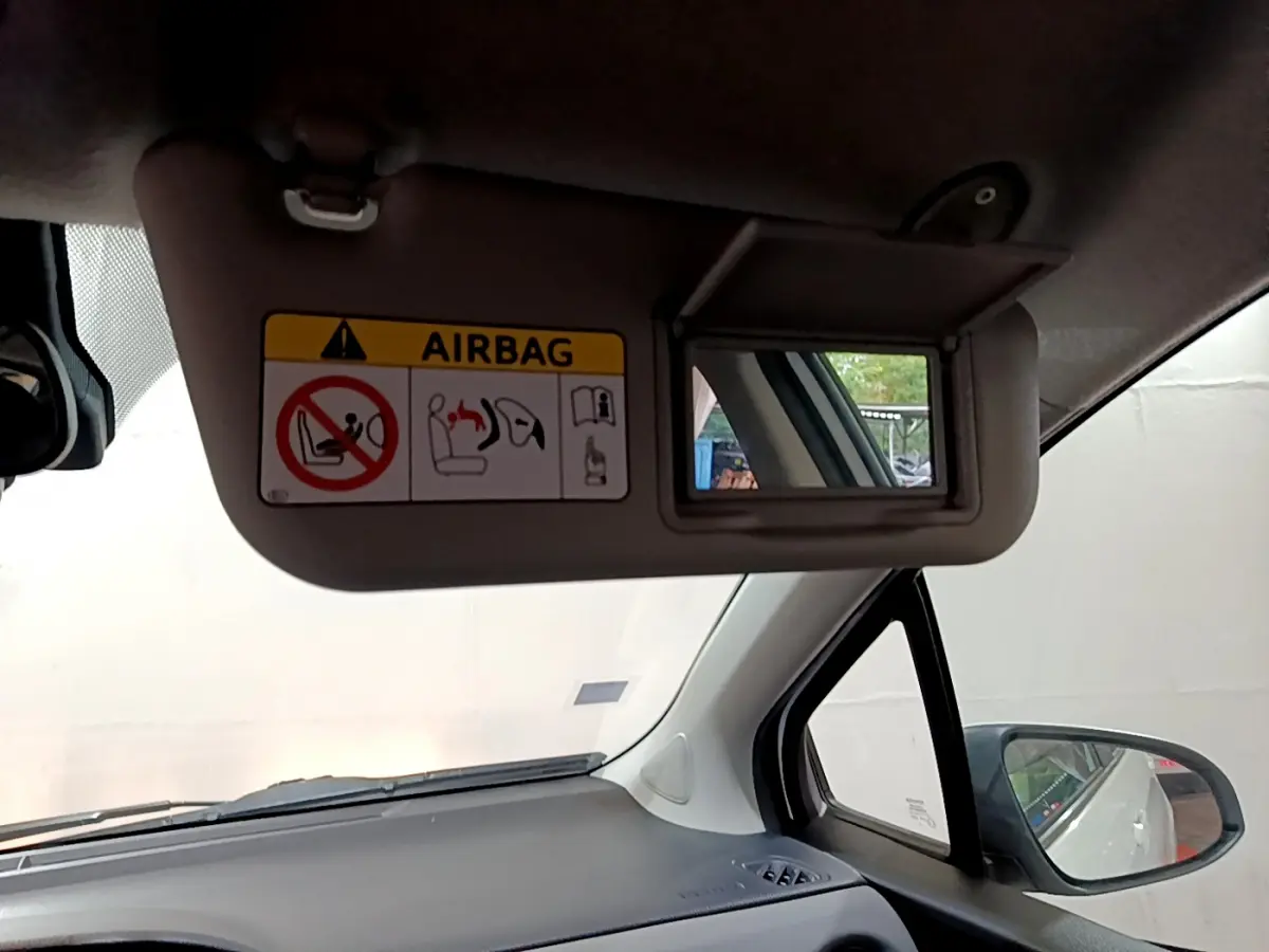 Miroir de pare-soleil côté conducteur avec avertissement airbag, tableau de bord gris et rétroviseur extérieur visible sur Toyota Yaris blanche.