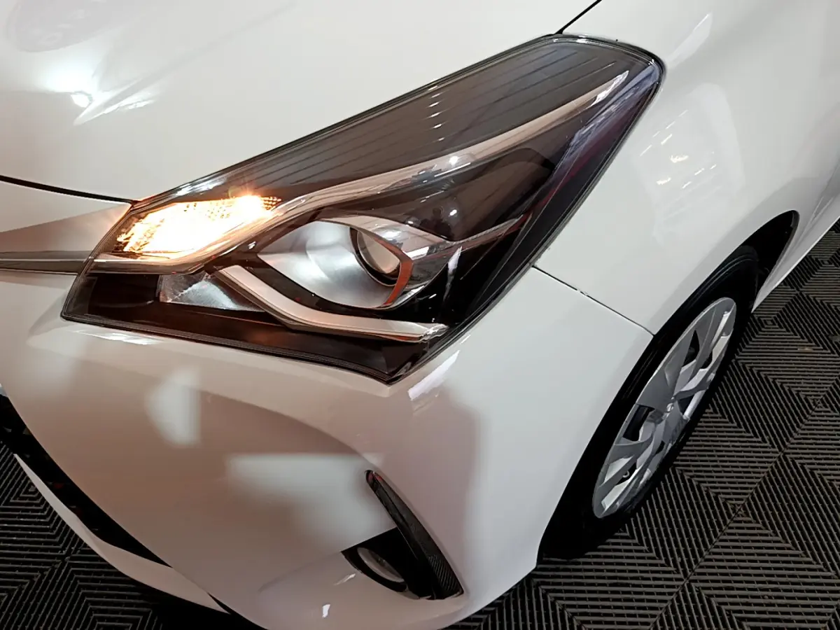 Gros plan sur le phare avant gauche allumé d'une Toyota Yaris blanche, avec partie de la roue visible.