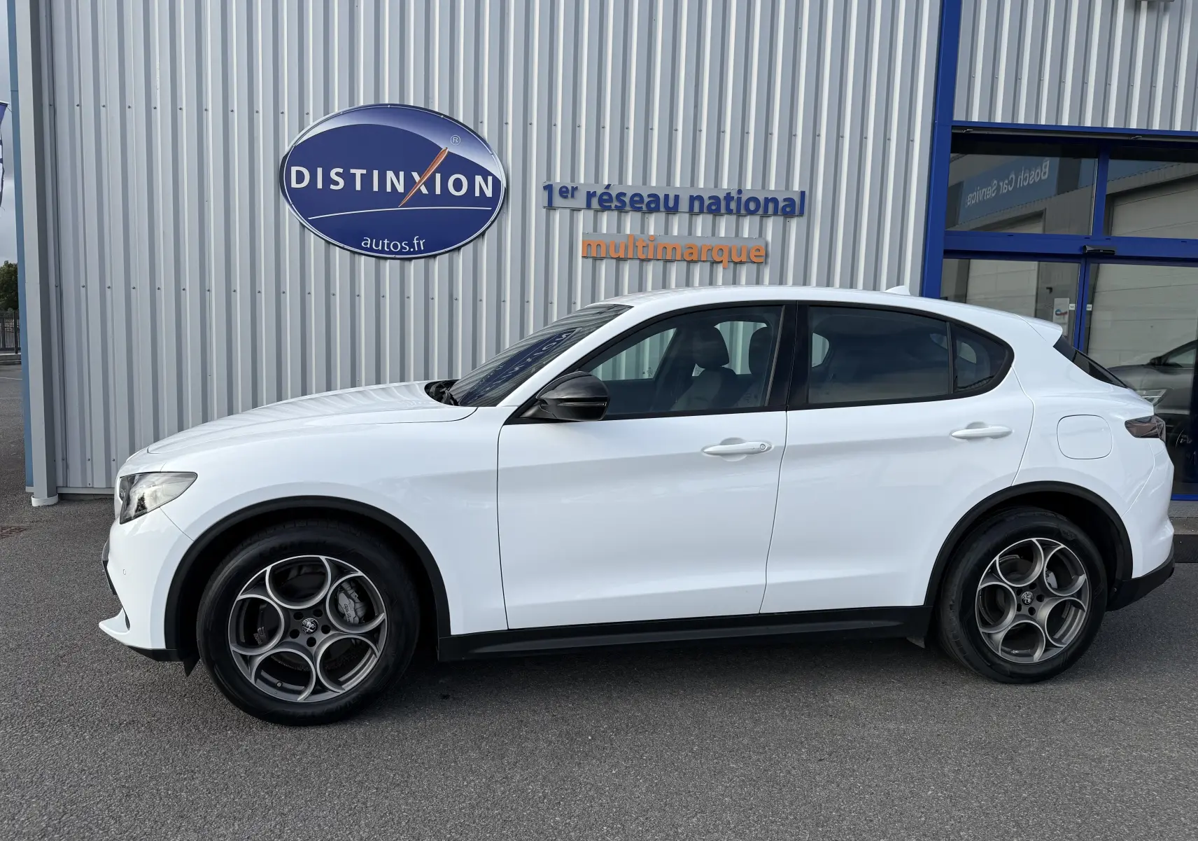 Profil côté droit de l'Alfa Romeo Stelvio blanc Alfa 2023, avec jantes spécifiques et rétroviseurs noirs, devant un bâtiment métallique.