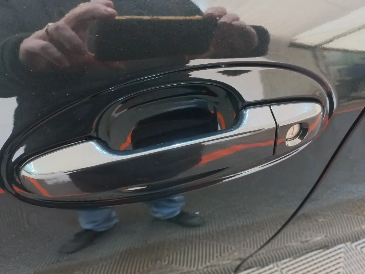 Poignée de porte avant droite chromée sur Toyota Yaris noire, reflet de la personne prenant la photo visible.