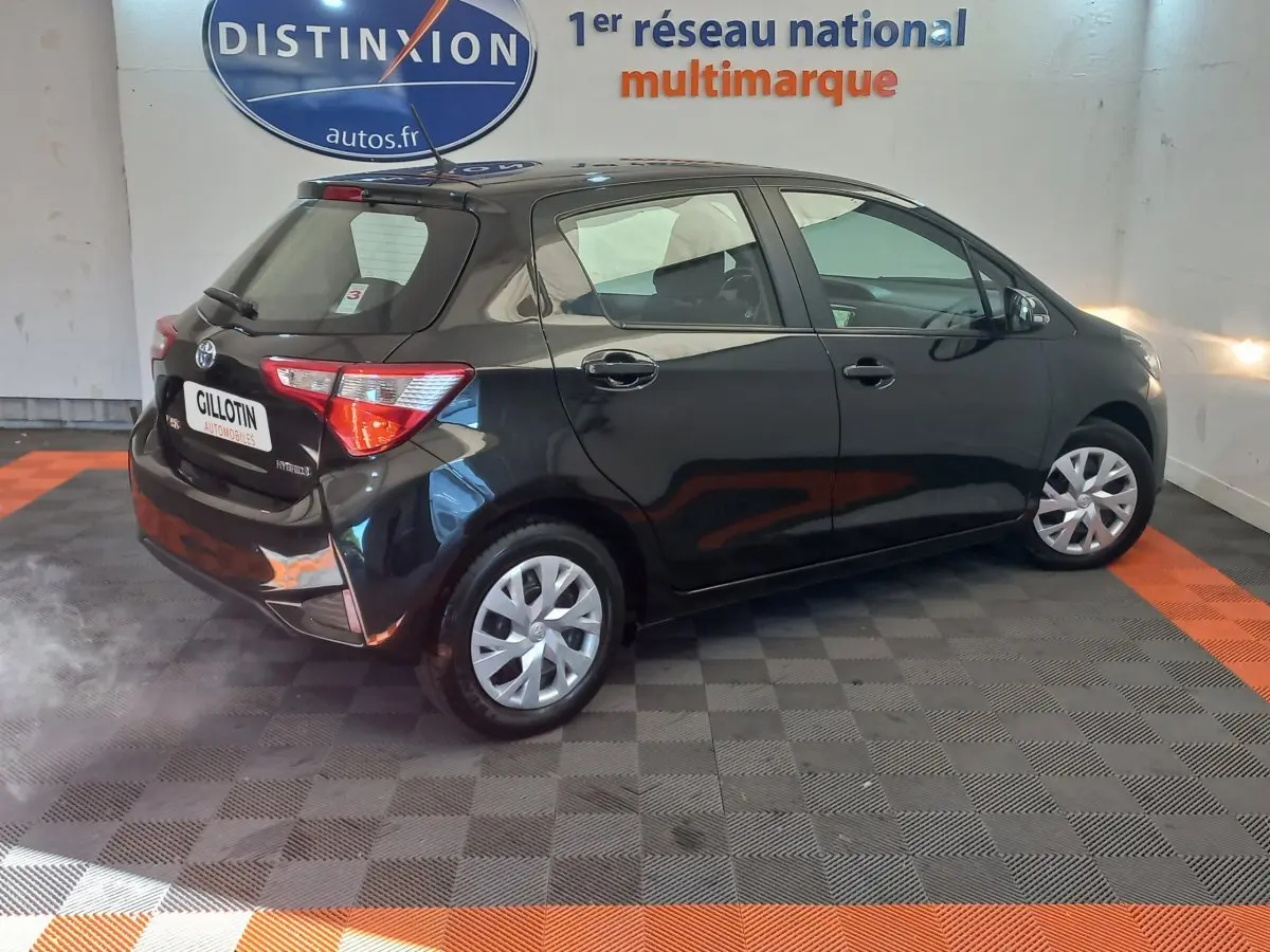 Toyota Yaris noire vue de trois quarts arrière côté droit dans un showroom avec sol gris et orange.