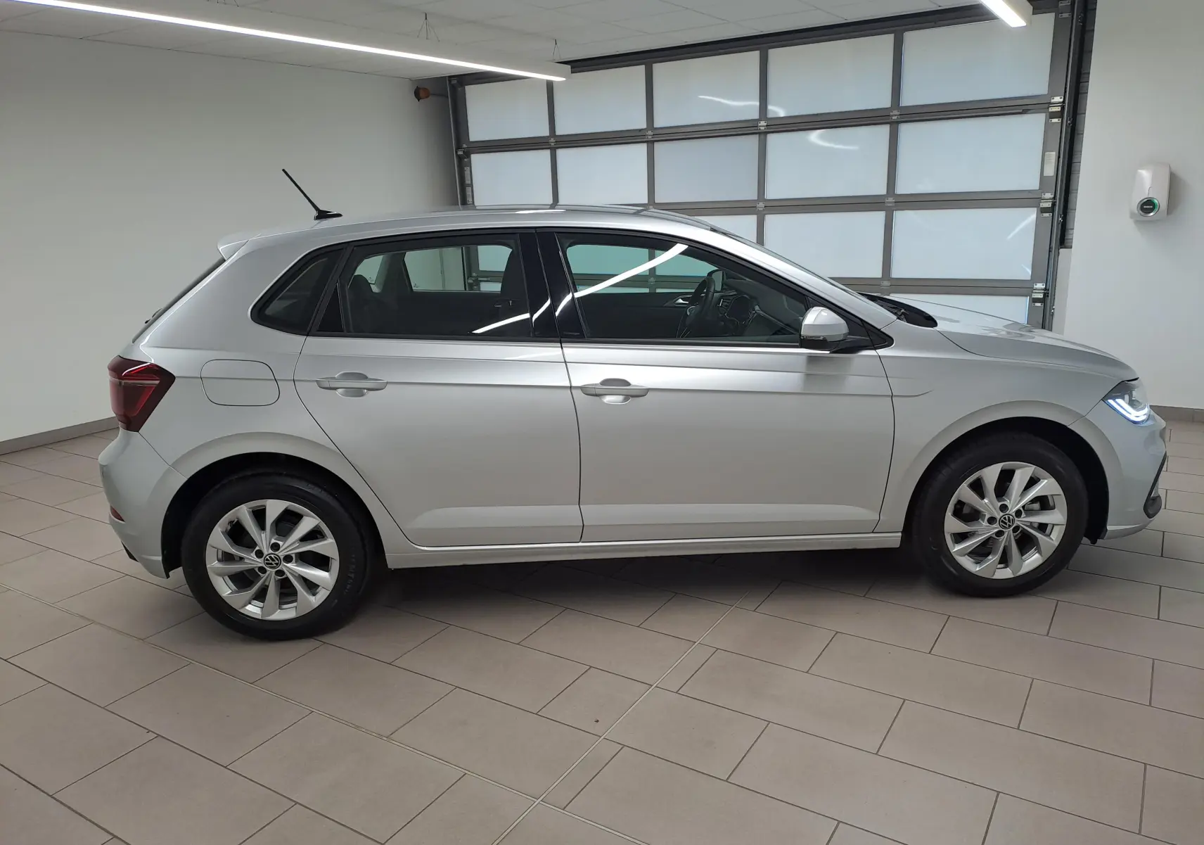 Vue de profil côté gauche d'une Volkswagen Polo 2025 en peinture reflet d'argent, avec feux avant allumés en intérieur showroom.