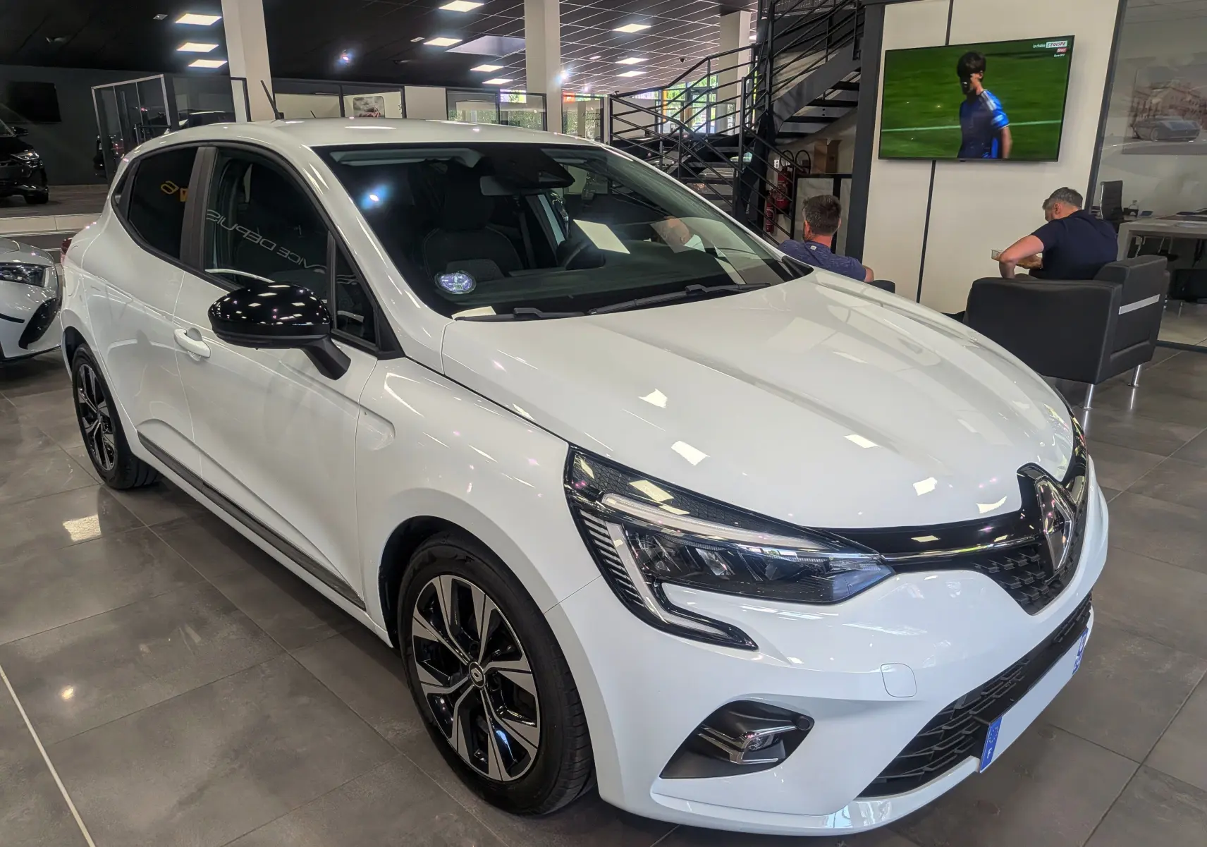Renault Clio V TCe 100 GPL blanc glacier vue 3/4 avant droit en intérieur avec jantes noires bicolores.