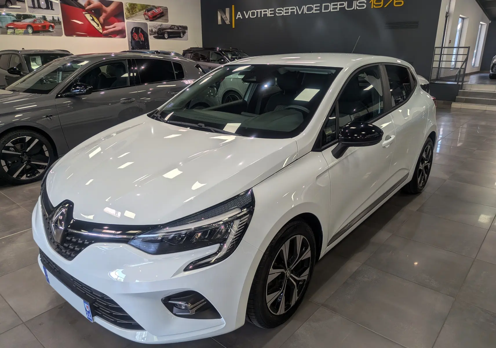 Renault Clio V TCe 100 GPL blanc glacier vue 3/4 avant droit en intérieur, jantes alliage bicolores visibles.