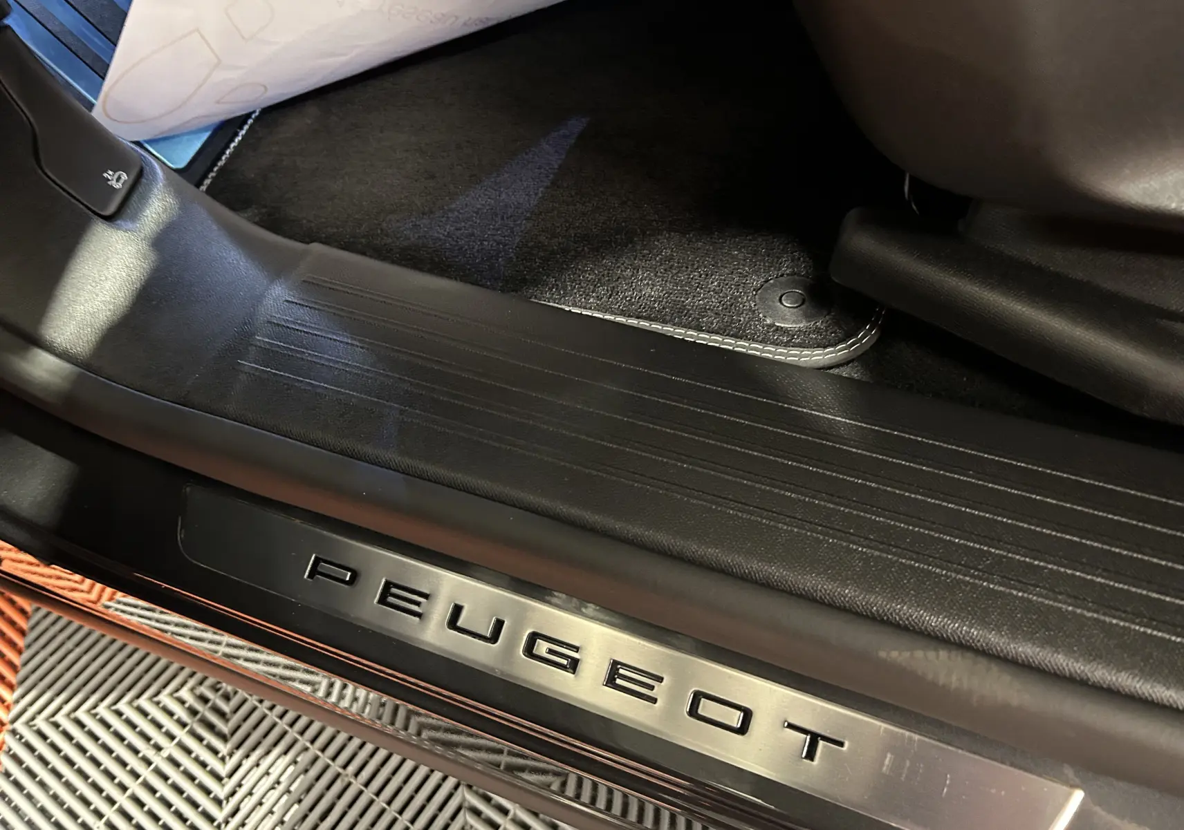 Gros plan sur la plaque de seuil de porte Peugeot avec intérieur noir, tapis de sol et partie de siège visible.