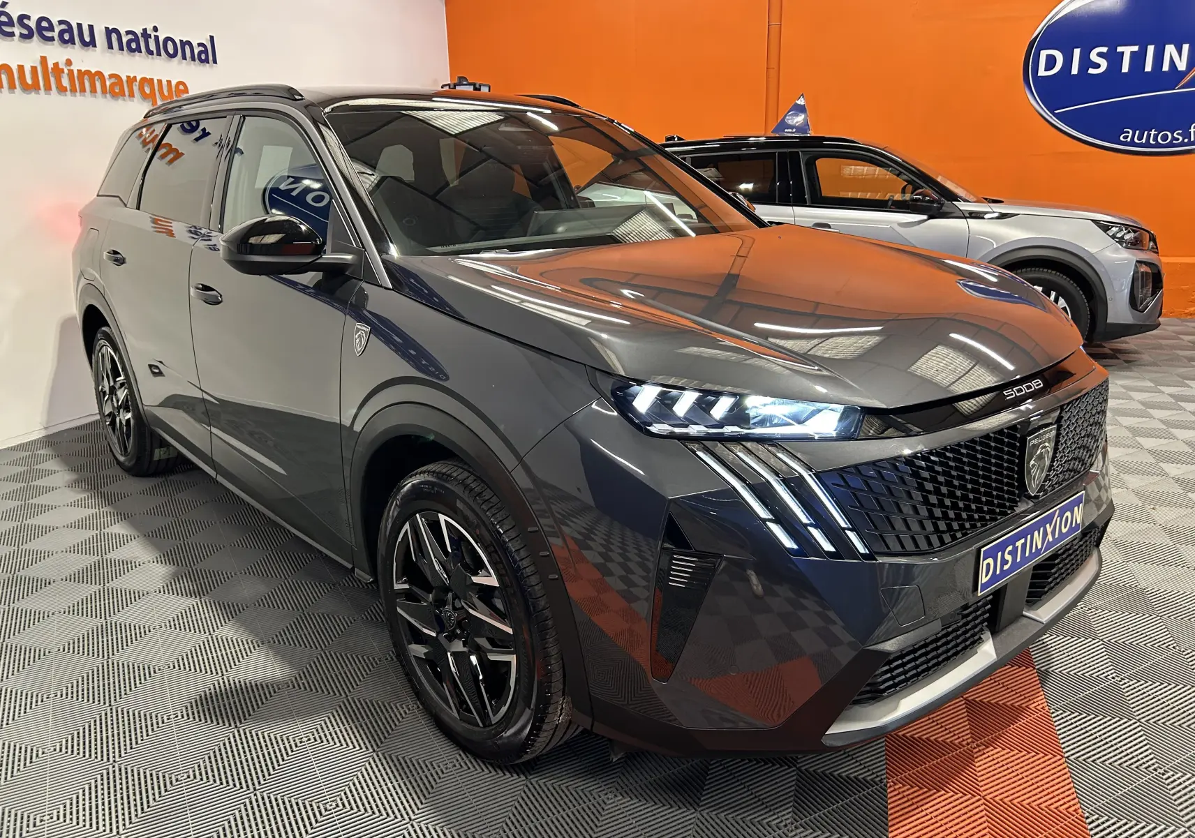 Peugeot 5008 Hybrid gris titane en 3/4 avant droit avec phares allumés dans un showroom orange.