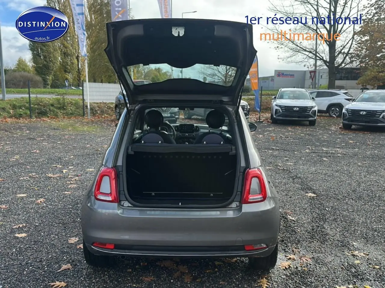 Vue arrière d'une FIAT 500 gris avec coffre ouvert, montrant l'intérieur et les sièges avant dans un parking extérieur.