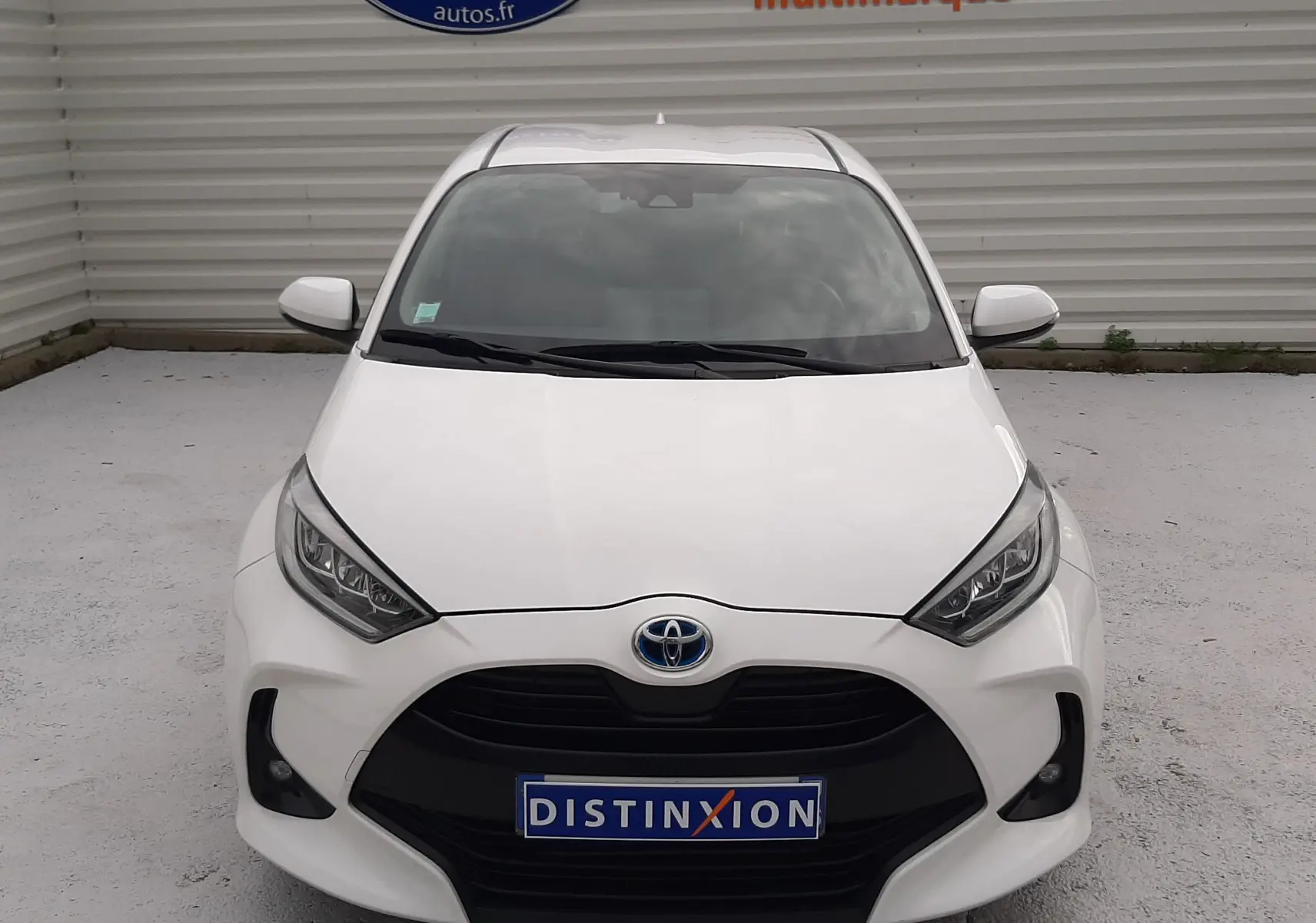 Vue frontale d'une Toyota Yaris blanche 2023 avec calandre noire et phares LED, stationnée devant un mur gris.