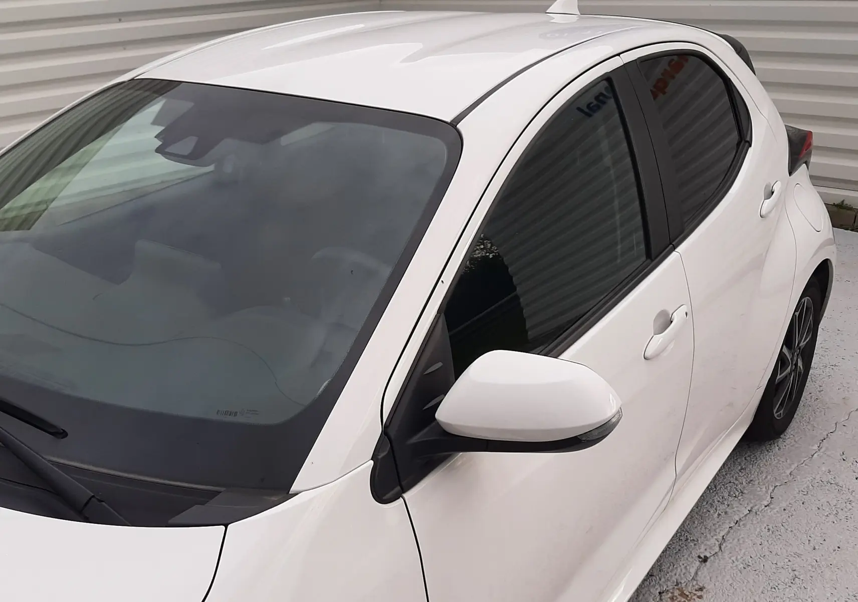 Toyota Yaris blanche vue 3/4 avant droit, avec vitres teintées et rétroviseur blanc sur fond gris.