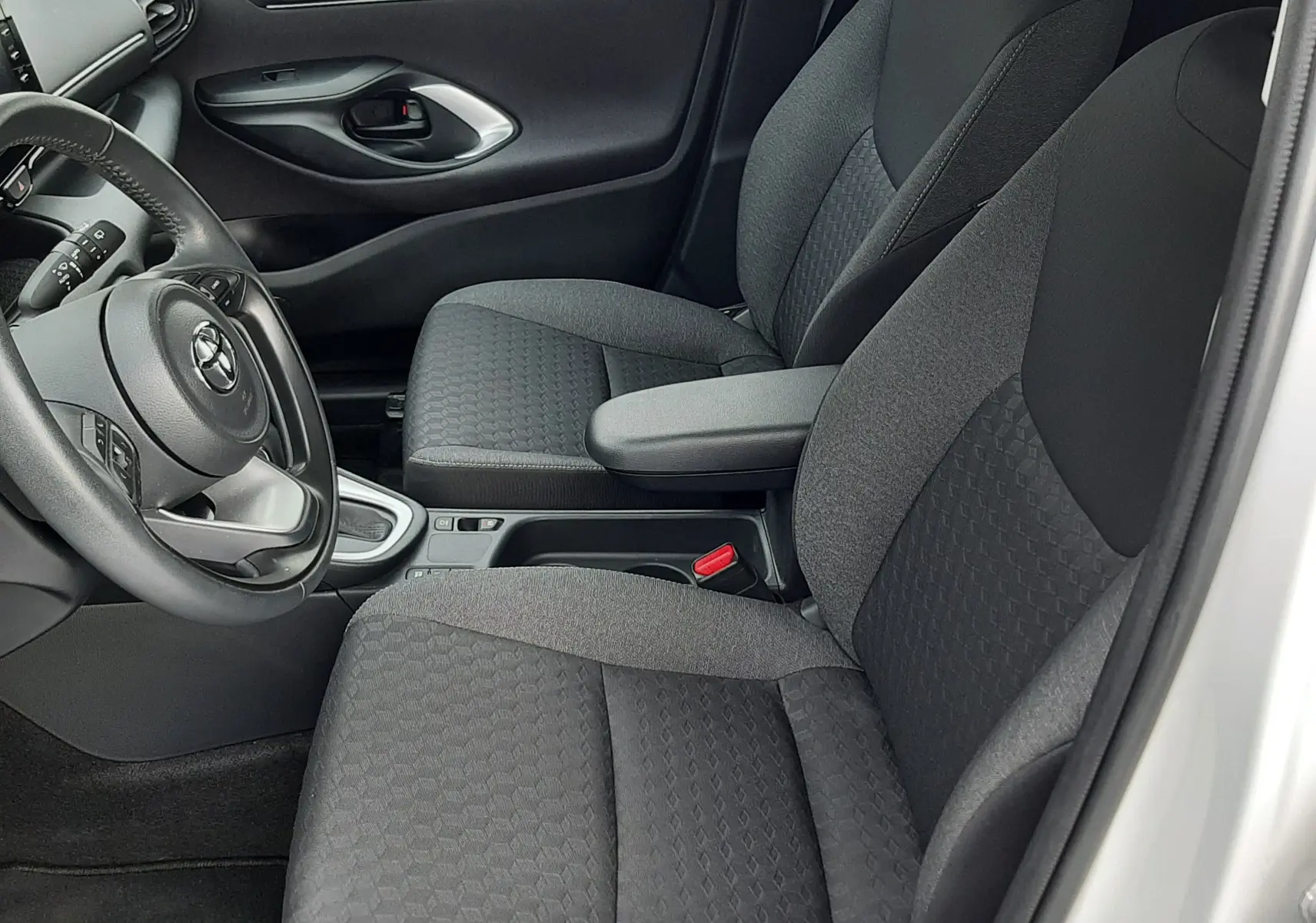 Vue intérieure côté conducteur de la Toyota Yaris blanche 2023, sièges tissu noir avec accoudoir central et volant multifonction.