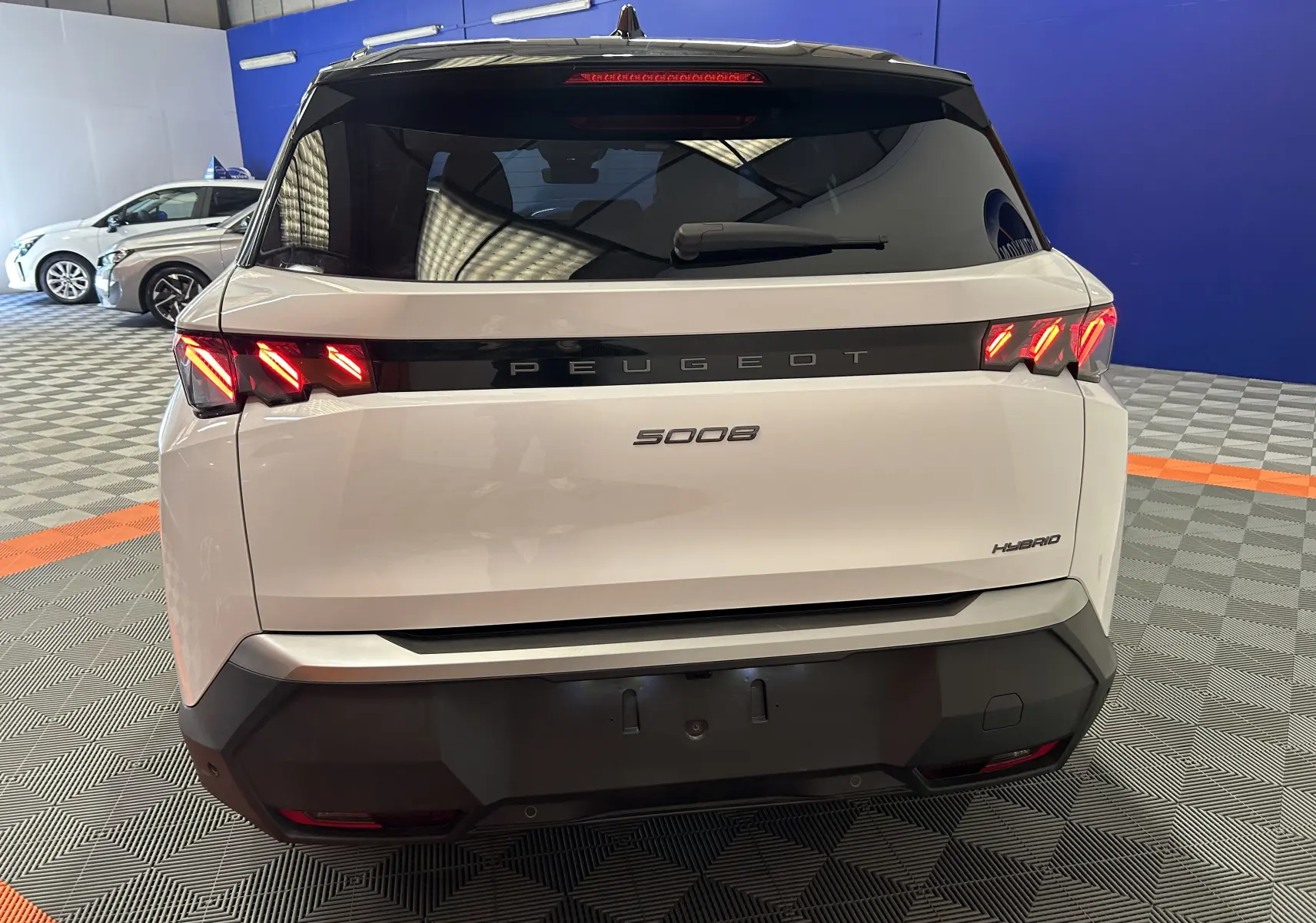 Vue arrière d’un Peugeot 5008 blanc hybride 2025 avec feux LED allumés et hayon électrique en intérieur showroom.