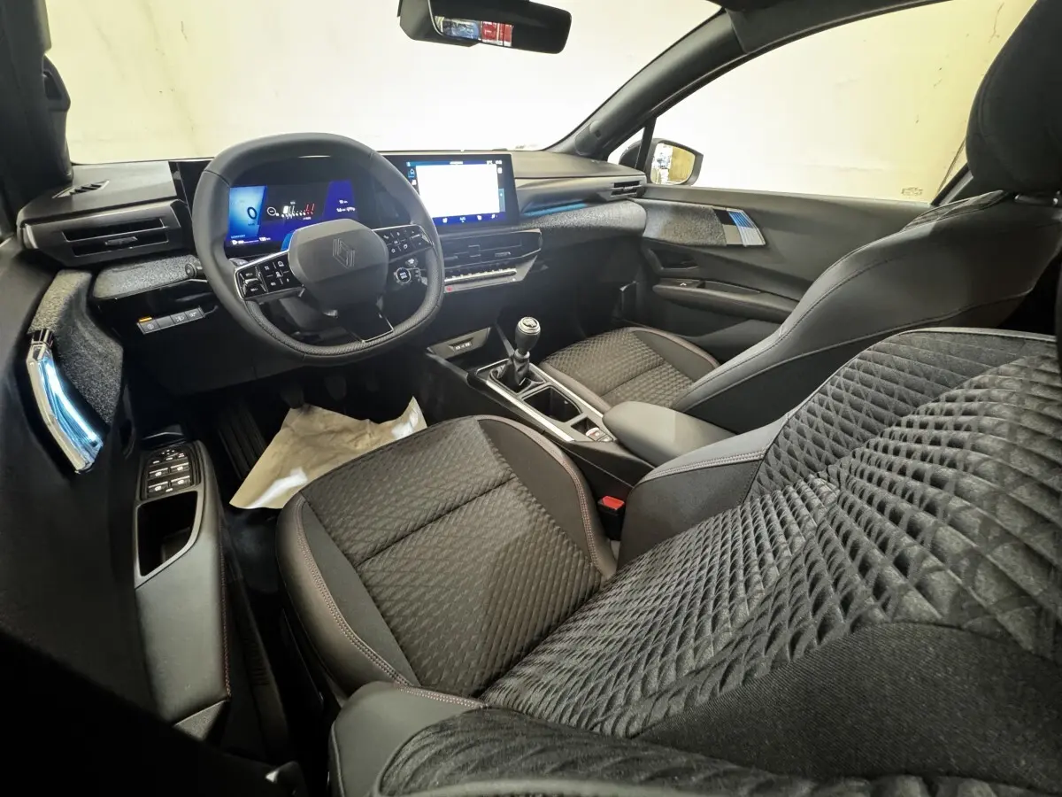 Vue intérieure avant droite de la Renault Clio Techno TCe 115 essence 2026 avec sièges noirs et tableau de bord numérique.
