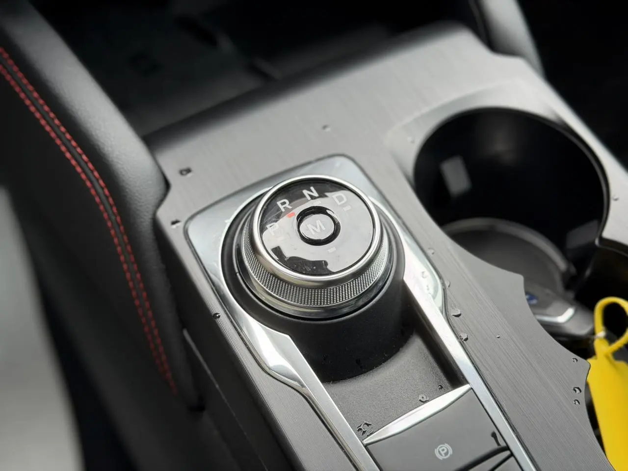 Gros plan sur le sélecteur rotatif de boîte automatique et accoudoir noir avec surpiqûres rouges dans un Ford Kuga noir Agathe.