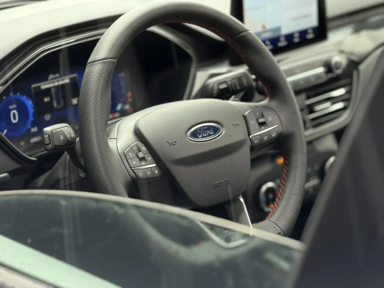 Vue rapprochée du volant noir perforé avec surpiqûres rouges du Ford Kuga 2023, tableau de bord et écran tactile visibles.
